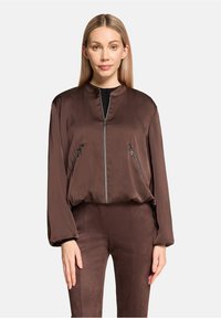 Brown bomber jacket with a high collar, front zipper, and two zippered side pockets. The material is smooth, with a relaxed fit and long sleeves.