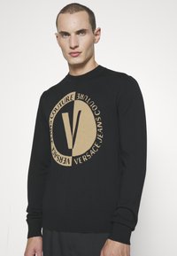 Zwarte gebreide trui met een ronde tan kleurige logo waarin de tekst "VERSACE JEANS COUTURE" en een prominente "V" in het midden zijn verwerkt. Geribde manchetten en onderrand.