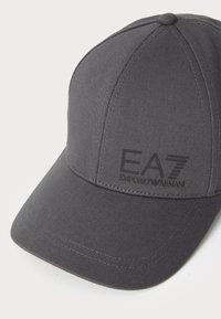 Gråt stoff baseballcaps med strukturert krone, buet bill og hevet svart logo på siden. Stingdetaljer er synlige.