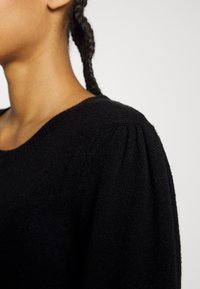 Pull en maille noire avec un col rond et des détails plissés aux épaules. Texture douce, conception ajustée, et bords côtelés.