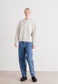Light beige sweatshirt with ribbed cuffs and hem, paired with loose-fitting blue jeans. Model wears black chunky boots. Plain background.