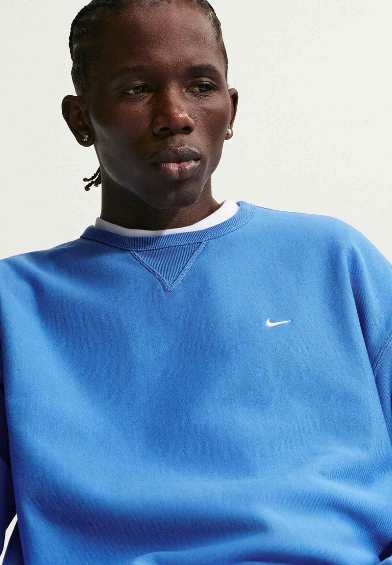 Junger Mann mit geflochtenem Haar, der einen blauen Nike-Sweatshirt trägt und nach unten schaut, vor einem schlichten, hellen Hintergrund.