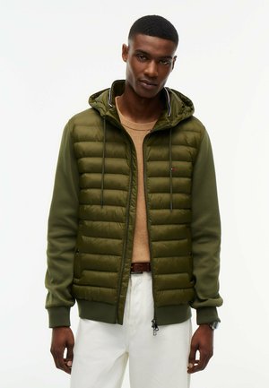 Mann trägt eine grüne Steppjacke mit Kapuze, einen braunen Pullover, weiße Hose und eine Uhr, steht vor einem einfarbigen hellen Hintergrund.