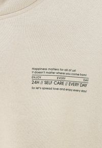 Der Text auf beige Stoff lautet: "Glück zählt für uns alle! Es spielt keine Rolle, woher du kommst! 24H // SELBSTFÜRSORGE // JEDEN TAG."