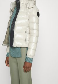 Person iført en blank cremet pufferjakke over en teal stribet skjorte og olivengrønne bukser, stående mod en simpel hvid baggrund.