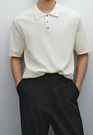 Homme portant un polo en maille à manches courtes de couleur crème, rentré dans un pantalon noir taille haute, les mains dans les poches.