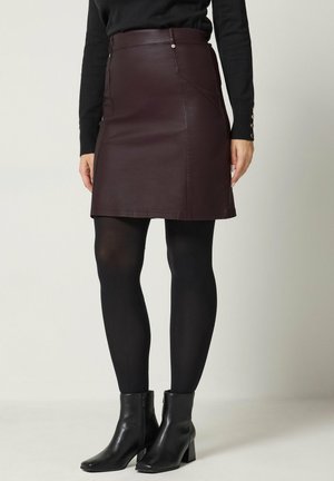 Donna che indossa una gonna di pelle marrone scuro, collant neri, stivaletti neri e un top nero a maniche lunghe con dettagli a bottoni sulle maniche.