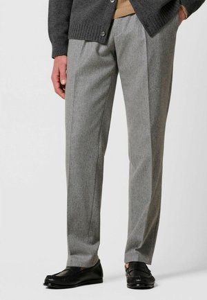 Mand iført grå skræddersyede bukser, sorte læder loafers, mørkegrå cardigan og tanfarvet sweater, står med hånden i lommen.