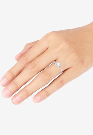 Anello in argento con una pietra preziosa rotonda e trasparente, montata su una semplice fascia. La pietra è circondata da accenti metallici decorativi.