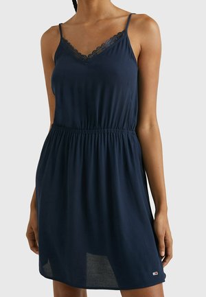 Frau trägt ein ärmelloses marineblaues Kleid mit Spitzenbesatz am Ausschnitt und einem elastischen Taillenband, steht vor einem einfarbigen Hintergrund.