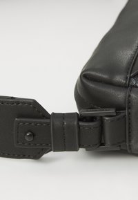 Colțul unei genți din piele neagră, prezentând o suprafață texturată, accesorii metalice și un detaliu cu curea cu cataramă, având o marcă discretă.