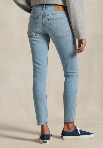 Asmuo dėvi šviesiai mėlynus siaurų kelnių džinsus, pilkšvą ilgomis rankovėmis palaidinę ir tamsiai mėlynas žemas sportinius batelius, stovintis prieš neutralią foną.