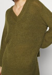 Personne portant un pull doux, olive vert, à manches longues avec un col en V et un tissu tricoté texturé, debout devant un fond uni.