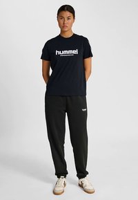 Czarna koszulka z krótkim rękawem z białym logo "hummel", w połączeniu z czarnymi dresami z nazwą marki. Model stoi z rękami na biodrach.