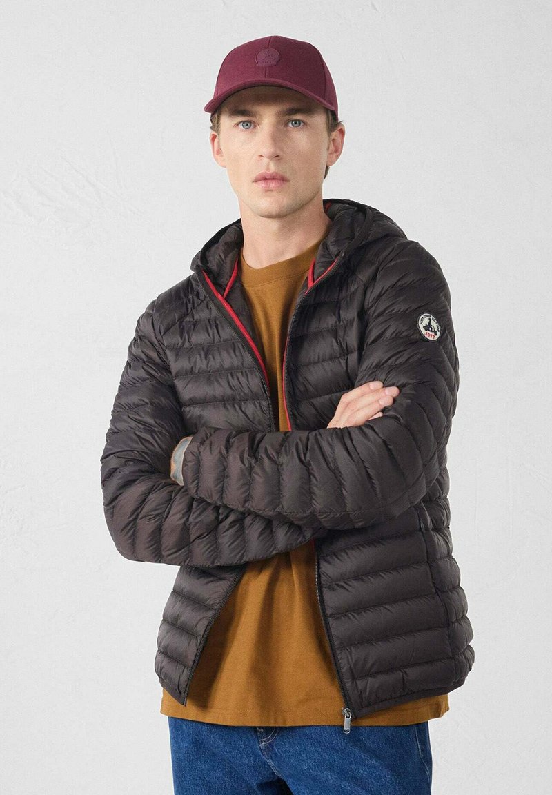 Giovane uomo con cappello bordeaux, camicia marrone e giacca trapuntata nera con cappuccio, braccia incrociate, in piedi davanti a uno sfondo bianco uniforme.