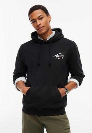 Giovane uomo che indossa una felpa nera Tommy sopra una camicia a righe con maniche rimboccate e pantaloni verdi, mani nelle tasche della felpa.