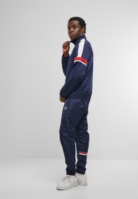 Navy tracksuit dengan kerah tinggi, aksen garis putih dan merah di lengan dan celana, manset elastis, dan detail logo di dada. Sneaker putih.
