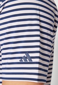 Dryžuota marškinėliai baltos ir tamsiai mėlynos spalvų, pagaminti iš minkštos medžiagos. Kairiajame rankove yra Adidas logotipas, atitinkančios spalvos.