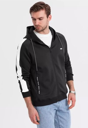 Sudadera con cremallera negra y rayas laterales blancas, con capucha ajustable. Cuenta con bolsillos con cremallera y marca a lo largo del ribete de los bolsillos. Tejido de mezcla de algodón.