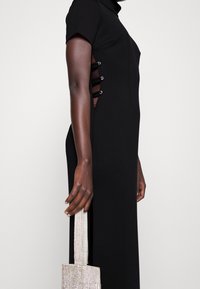 Musta mekko korkealla kauluksella ja lyhyillä hihoilla, jossa on sivuleikkauksia nappikoristeilla. Mallilla on kädess�ään karheatekoinen hopeinen käsilaukku.