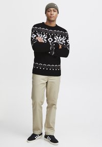 Black sweater with a white snowflake pattern, paired with tan pants and black sneakers. Model wears a green knit beanie, standing with arms crossed.