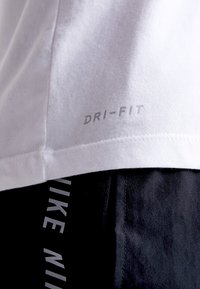 Camiseta Dri-Fit blanca con una textura suave, que presenta un logo impreso en gris claro. Tela negra con un logo de Nike en contraste debajo.