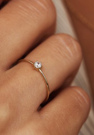 Goldring mit einem dünnen Band, das einen einzelnen runden klaren Edelstein in einer sechsflügeligen Fassung zeigt und auf einem Finger einer Person ruht.