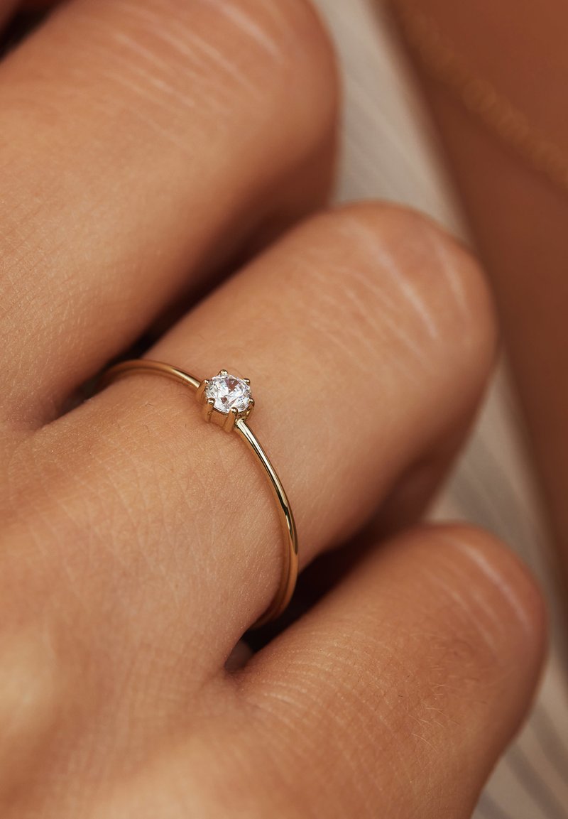 Goldring mit einem dünnen Band, das einen einzelnen runden klaren Edelstein in einer sechsflügeligen Fassung zeigt und auf einem Finger einer Person ruht.