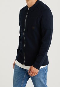 Marinblå zip-up jacka i texturerat tyg, med spetsig krage, bröstficka och slät dragkedja. Avslappnad passform med långa ärmar.