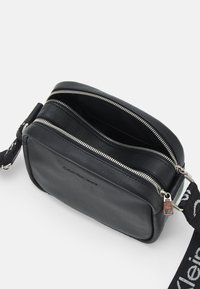 Borsa a tracolla in pelle nera di forma rettangolare, con due scomparti zip e una tracolla intrecciata che presenta il logo 'Calvin Klein Jeans'.