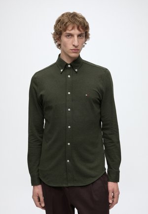 Jeune homme aux cheveux bouclés portant une chemise boutonnée vert foncé et un pantalon sombre, debout devant un fond clair uni.