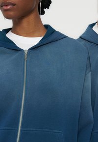 Κοντινό πλάνο δύο ατόμων που φοράνε μπλε φούτερ με φερμουάρ και λευκά πουκάμισα από μέσα, δείχνοντας την υφή του υφάσματος του φούτερ και το ασημένιο φερμουάρ.