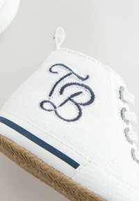 Baskets blanches en tissu texturé, accents bleus et logo embossé en bleu foncé. Elles présentent un devant à lacets et une semelle en caoutchouc à motif.