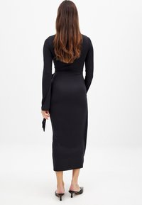 Black long-sleeve dress with a fitted silhouette; smooth, stretchy fabric; knee-length; low back design; paired with black heeled mules.