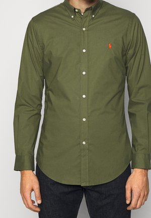 Homme portant une chemise kaki à manches longues boutonnée avec un petit logo rouge sur le côté gauche de la poitrine et un pantalon foncé.