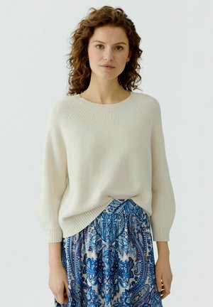 Femme aux cheveux bouclés portant un pull crème côtelé et une jupe taille haute à motif cachemire bleu et blanc, debout devant un fond blanc.