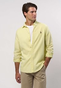 Mann trägt hellgelbes Hemd über weißem T-Shirt mit hochgekrempelten Ärmeln, beigefarbene Hose, steht mit einer Hand in der Tasche vor weißem Hintergrund.