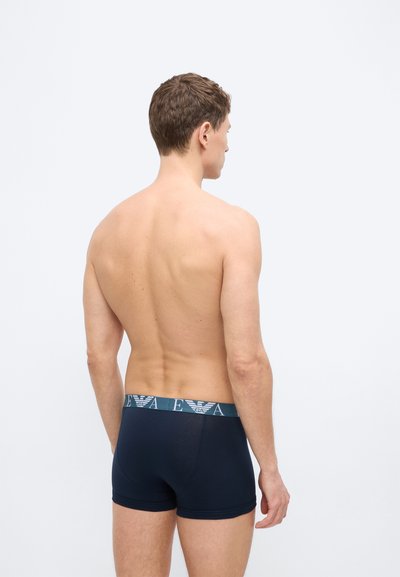 Άνδρας χωρίς πουκάμισο, στέκεται με την πλάτη προς τα εμπρός, φορώντας σκούρα μπλε βρακάκια boxer με λογότυπο στη μέση, σε απλό φωτισμένο φόντο.