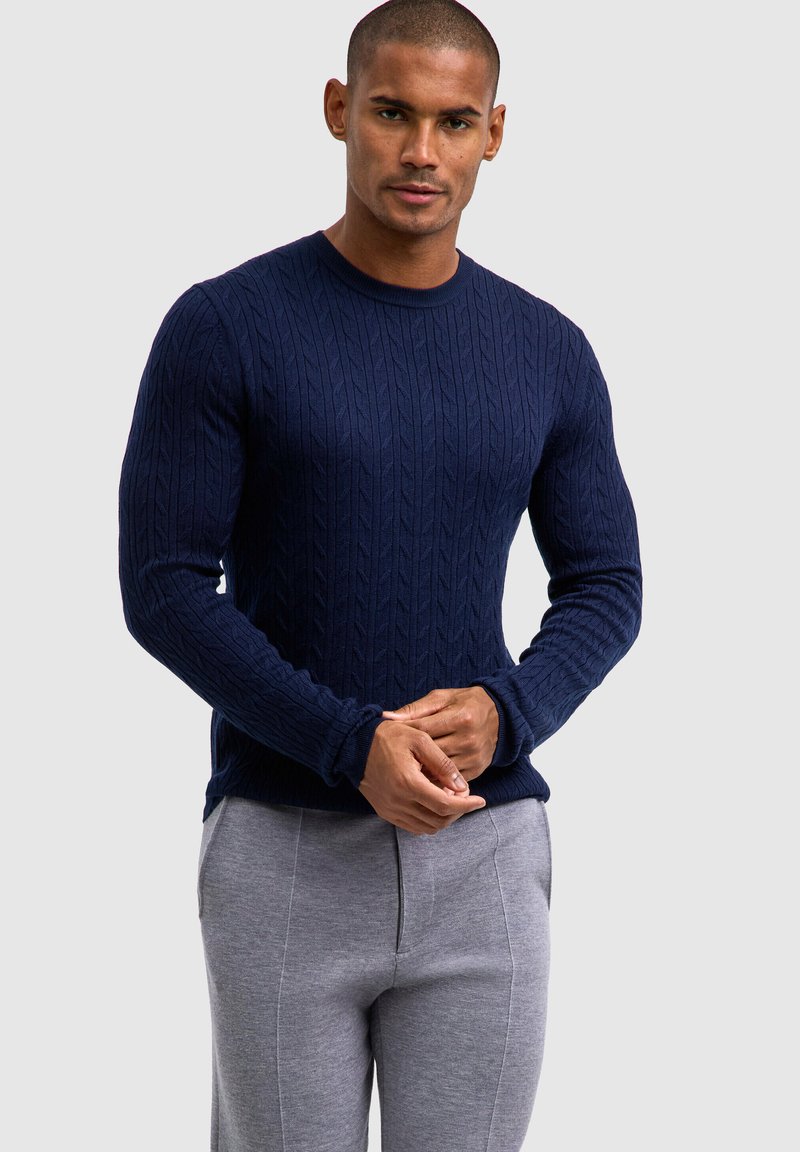 Granatowy sweter z dzianiny o wzorze w warkocz, okrągły dekolt i długie rękawy, w połączeniu z szarymi spodniami z detalem środkowego szwu.