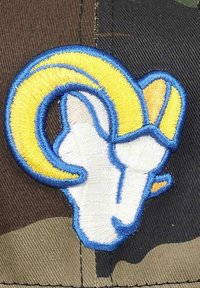 Gesticktes Abzeichen mit einem Widderkopf in Weiß und Gelb, umrandet in Blau, auf einem Camouflage-Stoffhintergrund.