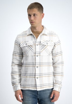 Man wearing white and beige plaid button-up shirt with front pockets, paired with blue jeans, looking slightly to the side on plain background.