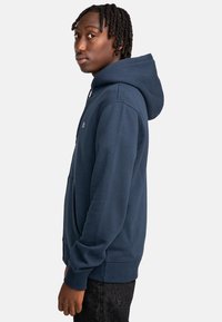 Marineblauer Kapuzenpullover aus weichem Stoff, mit einer Fronttasche, gerippten Bündchen und einem kleinen Logo auf der Brust.