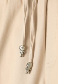 Beżowa tkanina z dwoma wiszącymi sznureczkami, z których każdy zakończony jest małym błyszczącym srebrnym zawieszkiem w stylu kreskówkowej pszczółki.