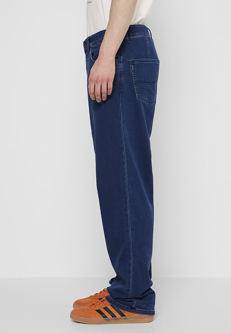 Jeans en denim bleu foncé avec une coupe droite, comportant une poche arrière et une taille standard. Associés à des chaussures de sport orange.