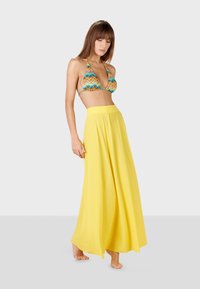 Falda maxi amarilla, de talle alto y fluida, hecha de una tela ligera. Combinada con un top de bikini de patrón zigzag multicolor.