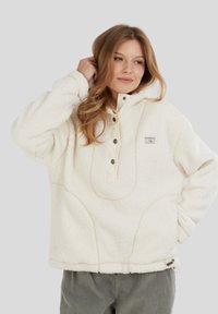 Kvinde iført en hyggelig hvid sherpa-pullover med trykknapper, stående med den ene hånd bag hovedet og den anden på hoften.