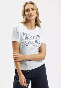 Jasnoniebieska bawełniana koszulka z białą kieszonką w kwiatowy wzór i napisem "Cross Jeans". Krótkie rękawy, luźny krój, gładka tekstura.