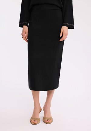 Kobieta ubrana w czarną midi ołówkową spódnicę i beżowe sandały na obcasie z odkrytymi palcami, stojąca na jednolitym jasnym tle.