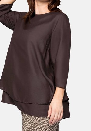 Lekka, ciemnobrązowa bluzka z długim rękawem, o zakrzywionym dołu i warstwowej konstrukcji. Gładka faktura i luźny krój. Poniżej wzorzyste spodnie.