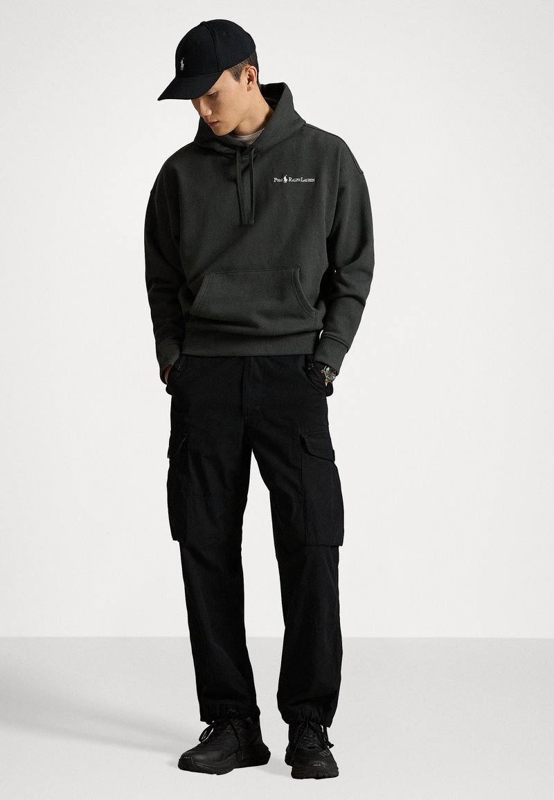 Dunkelgrüner Hoodie mit einer Vorderkante und Logo auf der Brust, kombiniert mit schwarzen Cargo-Hosen, die mehrere Taschen haben, und schwarzen Sneakers.